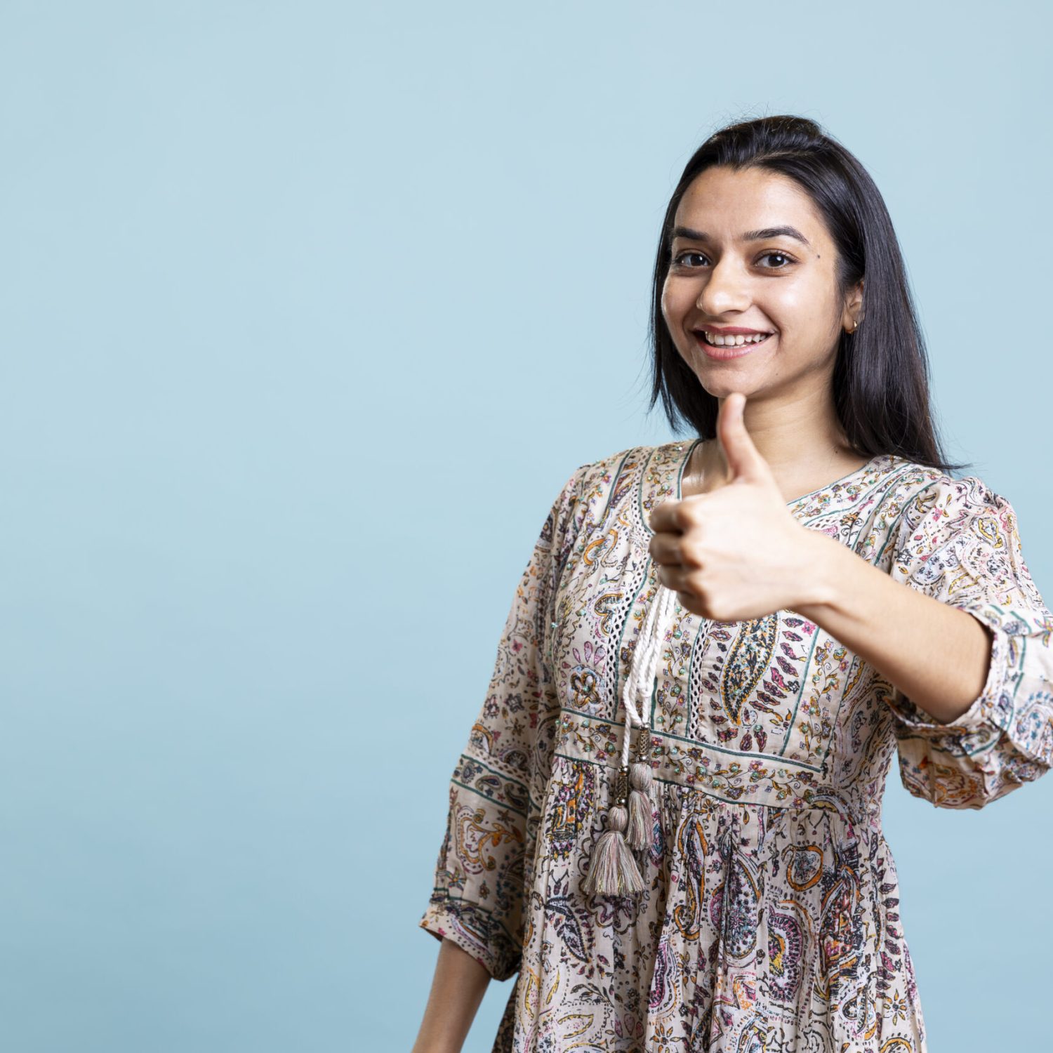 Joyful amazed person gives thumbs up okay symbol on camera, acting relaxed and positive against blue background. Young indian woman being optimistic and showing satisfaction with sign.